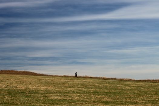 A lone person walks across an expansive grassy meadow beneath a cloudy sky, evoking solitude and calm.