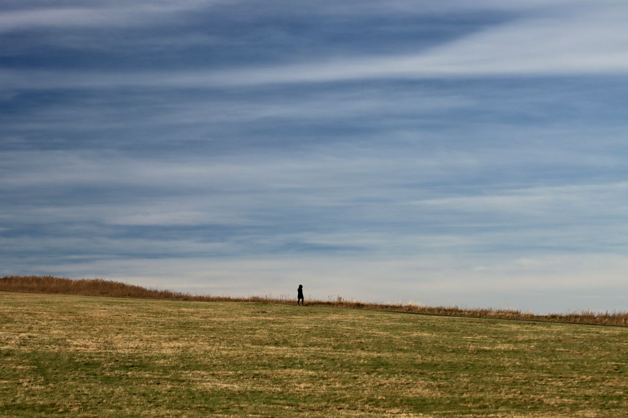 A lone person walks across an expansive grassy meadow beneath a cloudy sky, evoking solitude and calm.