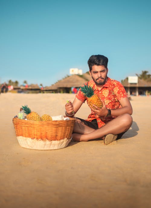 Photo of Man Sitting on Sand with Fruit Basket in front of Him Holding a Pineapple