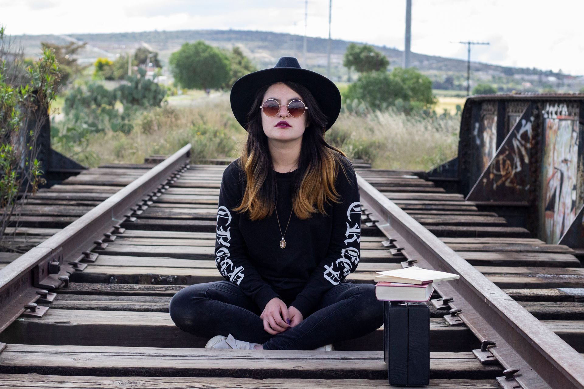 Stylish woman in black attire sitting on railroad tracks with books and suitcase nearby.