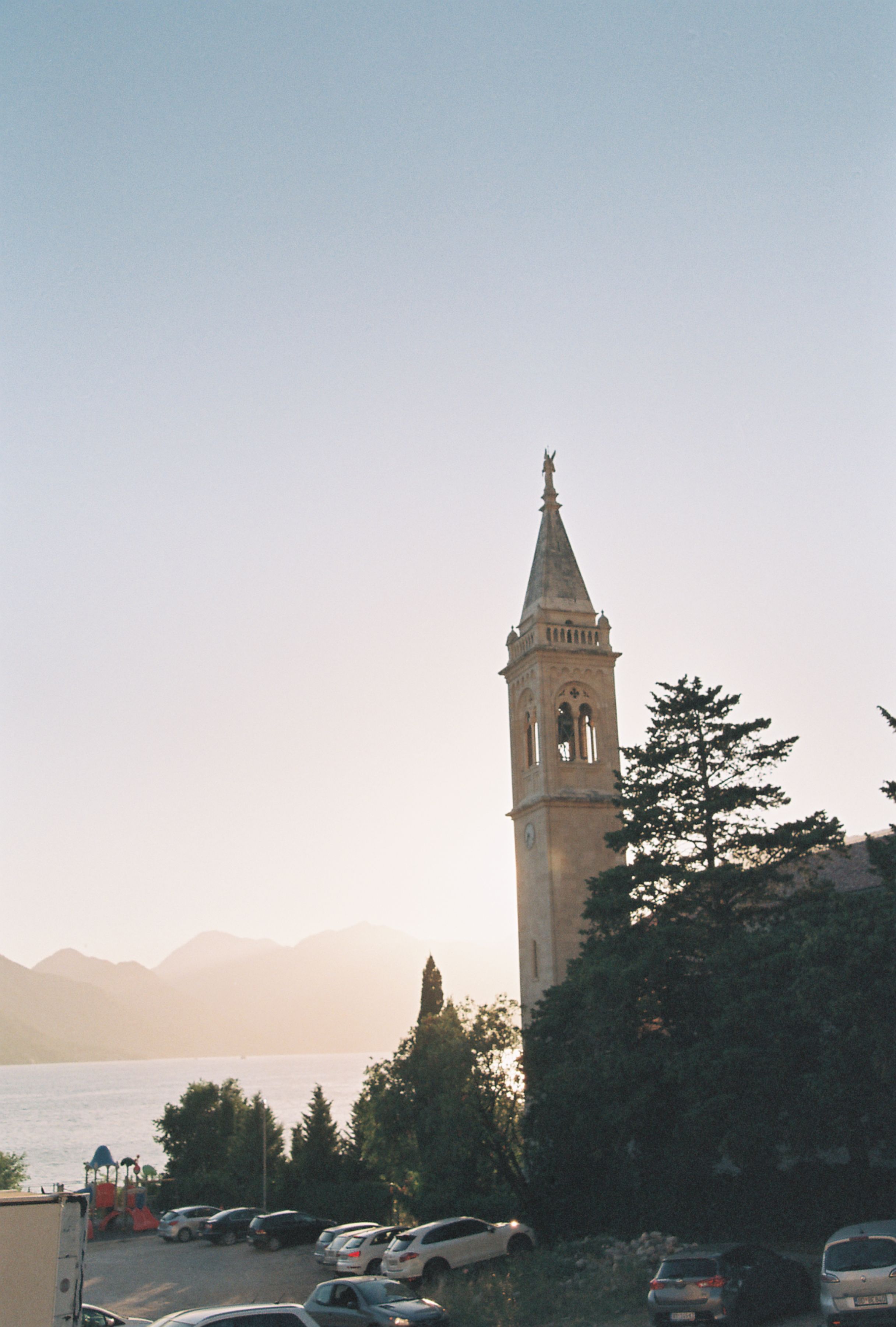 A serene view of a church tower at sunset in Kotor, Montenegro, with mountains and bay in the background.