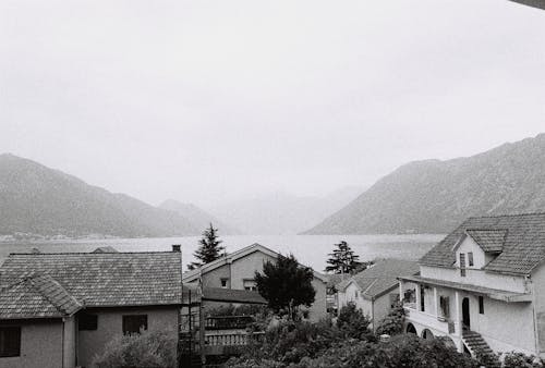 Black and White Photo of Houses by a Body of Water in Mountains 