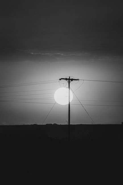 Black and white photo of the sun setting behind power lines, creating a dramatic silhouette.
