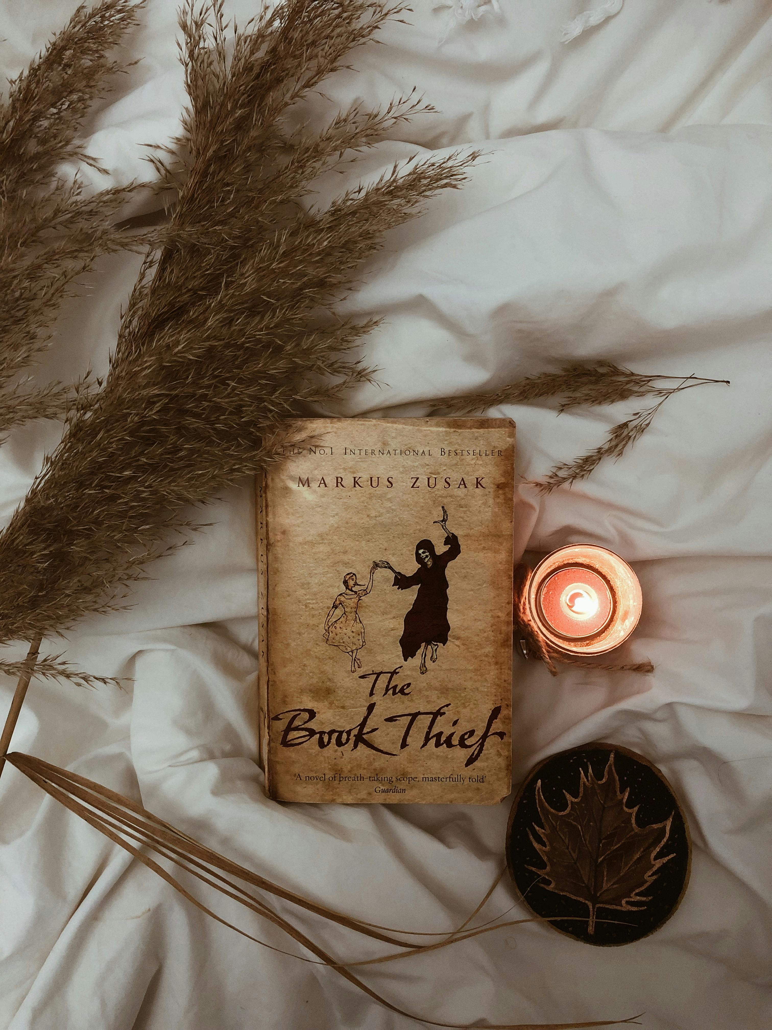 A cozy flat lay featuring 'The Book Thief' book surrounded by candles and dried plants on white fabric.