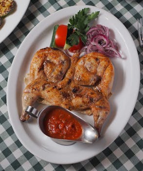 Appetizing grilled chicken with dipping sauce on a white plate, accompanied by vegetables.