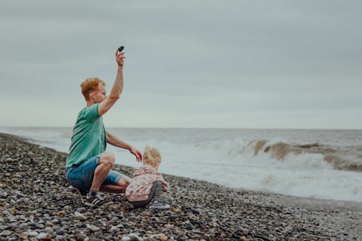 A father crouches with his daughter on a pebble beach, enjoying the overcast seashore.