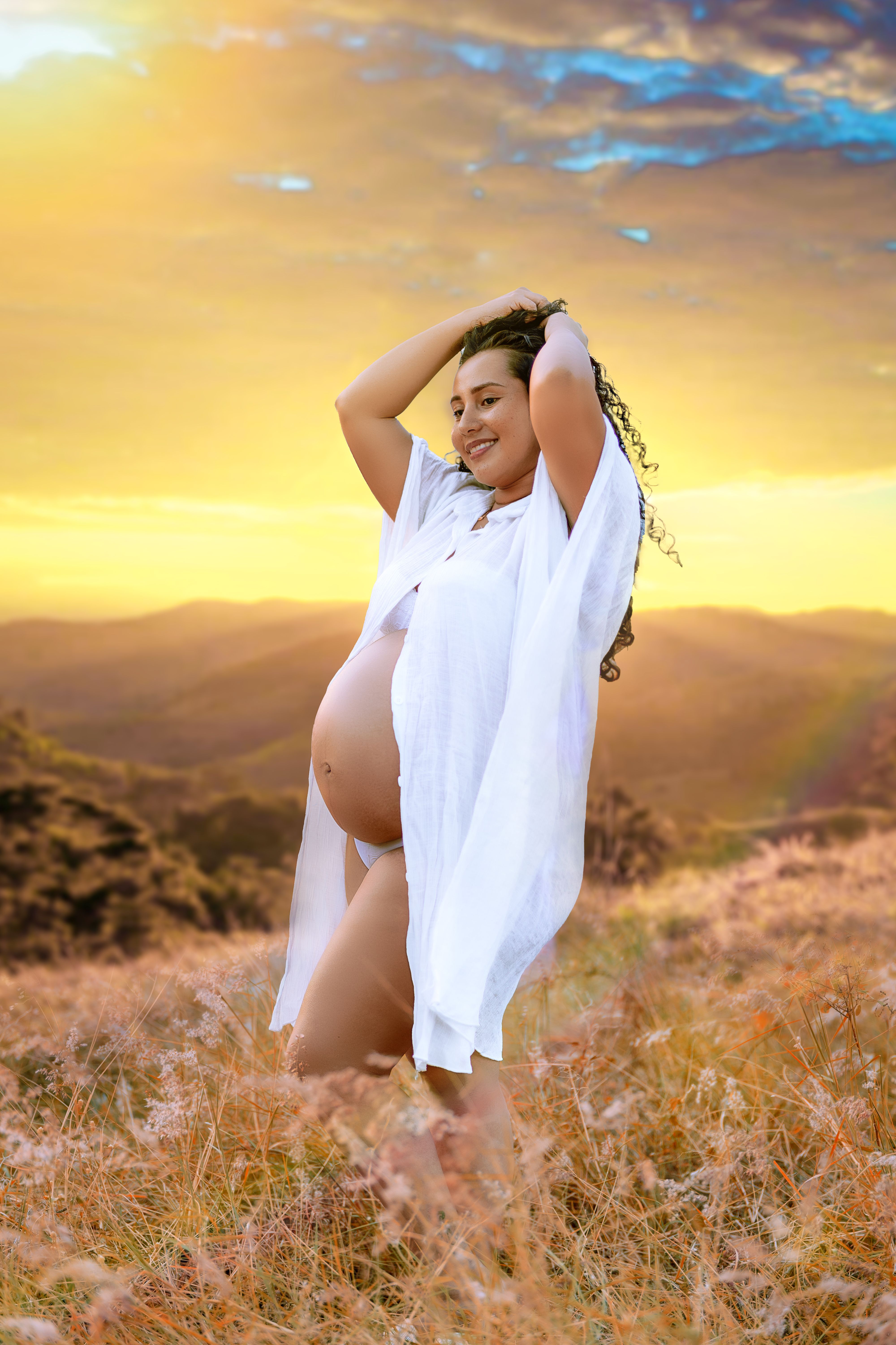 Pregnant woman joyfully standing in a scenic field during sunset, capturing happiness and beauty.