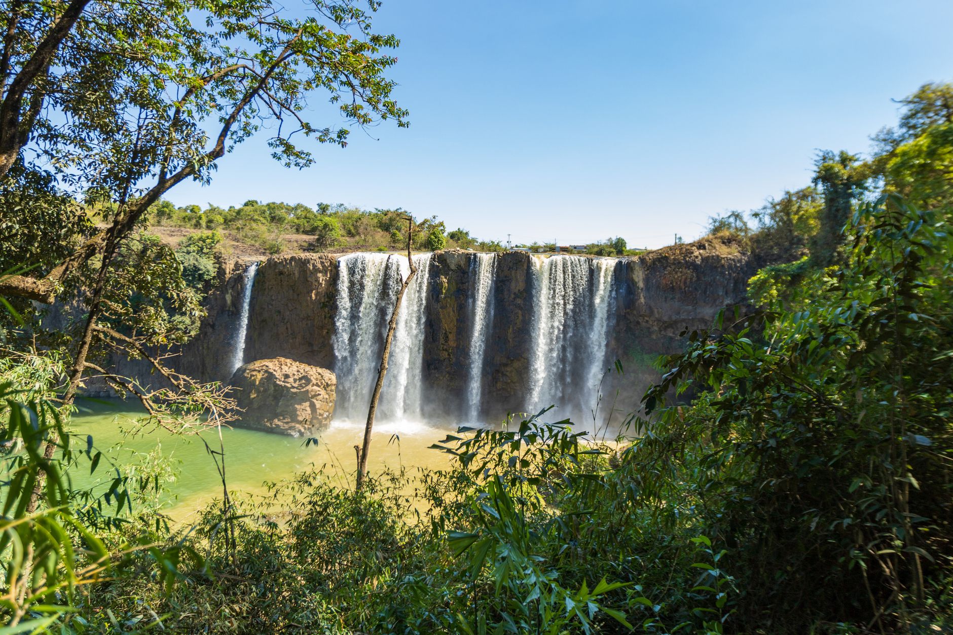 A mesmerizing waterfall cascading in Lâm Đồng, Vietnam, surrounded by lush vegetation and blue skies.