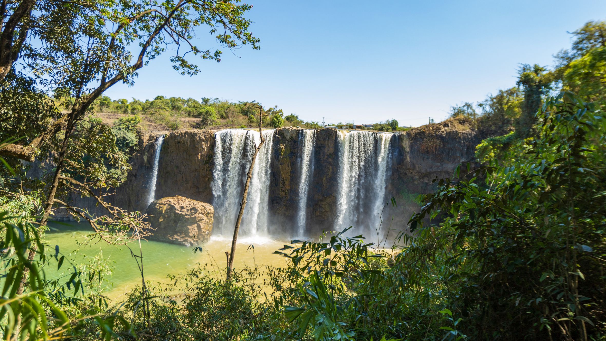 A mesmerizing waterfall cascading in Lâm Đồng, Vietnam, surrounded by lush vegetation and blue skies.