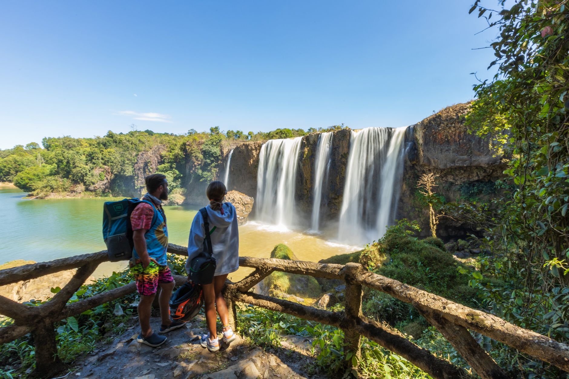 Two travelers enjoy the scenic view of Dray Nur Waterfall on a sunny day in Vietnam's Lâm Đồng province.