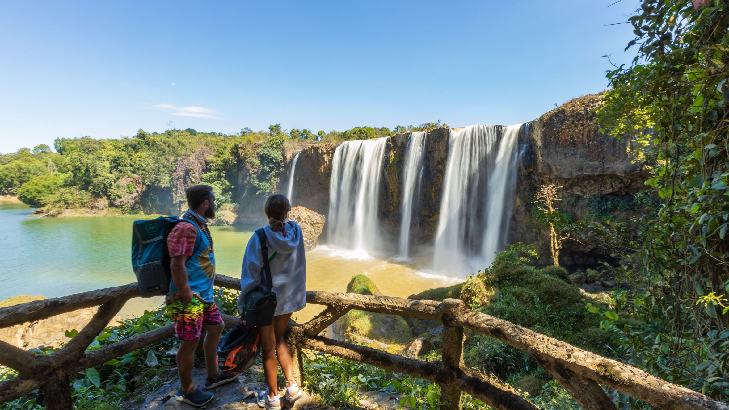Two travelers enjoy the scenic view of Dray Nur Waterfall on a sunny day in Vietnam's Lâm Đồng province.