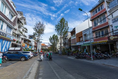 Free Urban street with motorbikes and cars, lined with shops and trees under a clear blue sky. Stock Photo