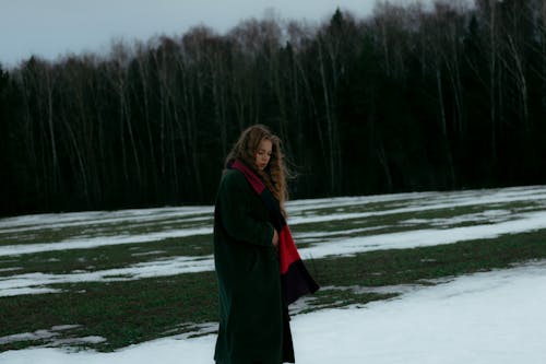 Young Woman in a Coat and Scarf Standing on a Field near Trees 