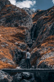 A man standing on a bridge in a rocky canyon with a waterfall, showcasing rugged nature.
