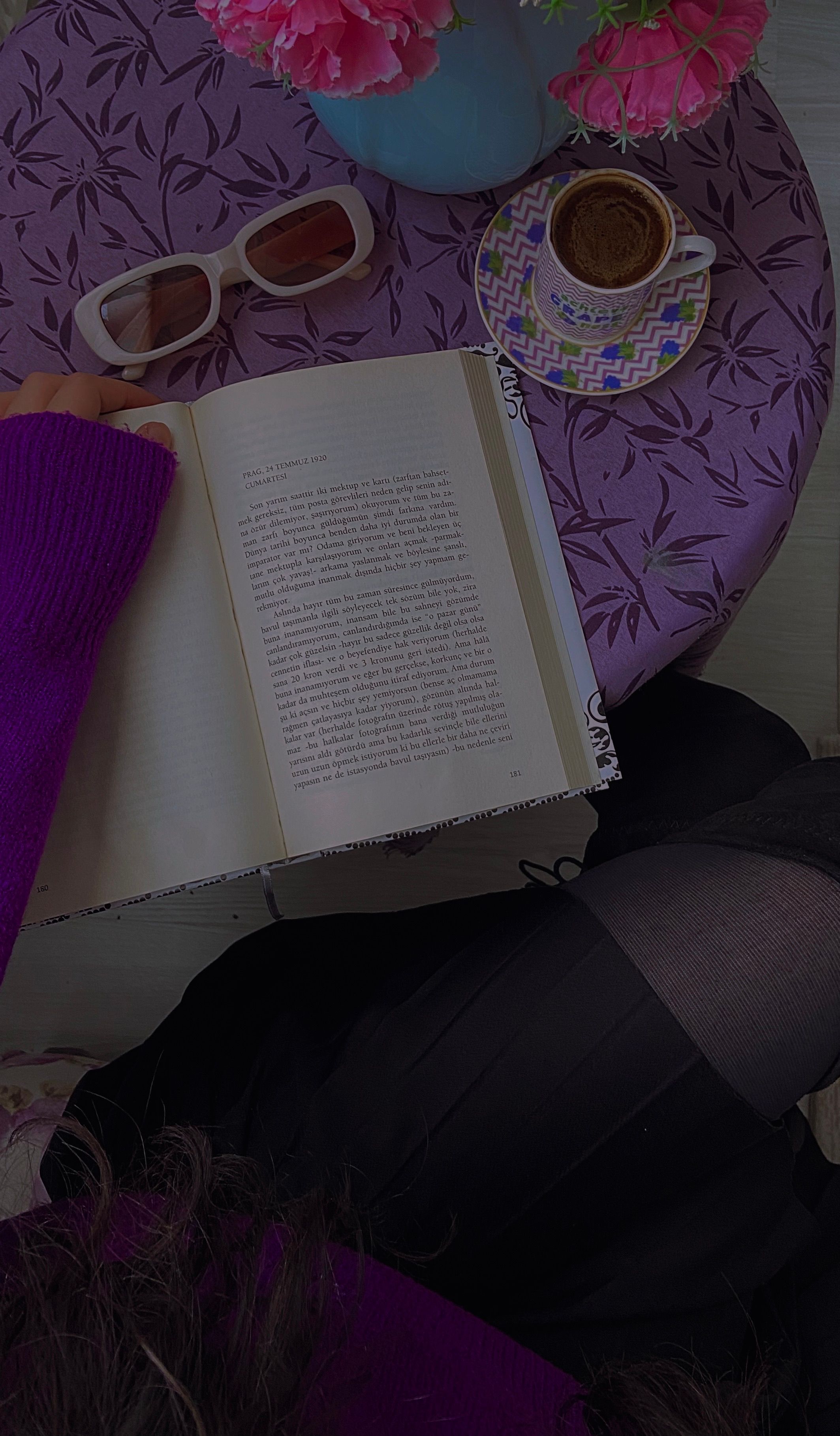 A high-angle view of a book with coffee, flowers, and sunglasses on a purple tablecloth.