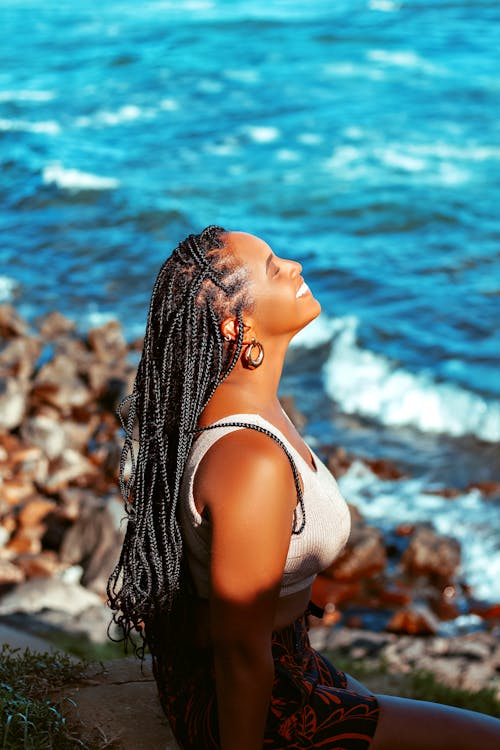 A Side View of a Braided Haired Woman in White Tank Top Sitting Near the Beach