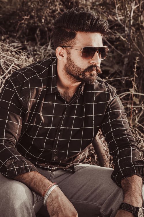 Free Stylish bearded man in sunglasses wearing a checkered shirt, seated outdoors. Stock Photo