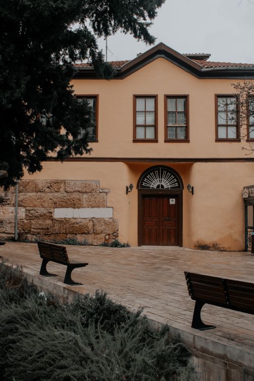 Free Historic beige building with wooden details, benches, and tree shadows in a tranquil outdoor setting. Stock Photo