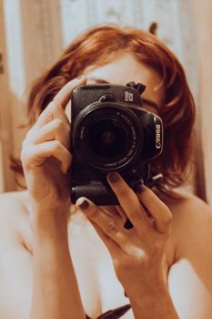 Redhead woman capturing her reflection in a mirror with a camera, indoor selfie.