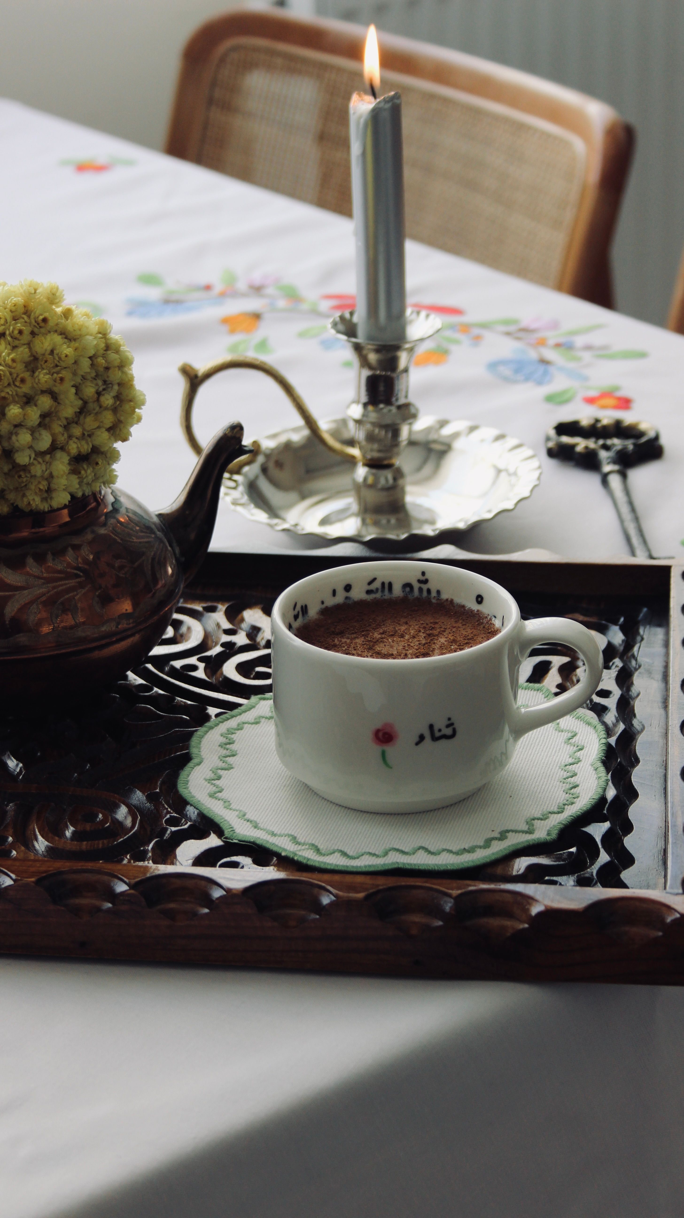 A cozy coffee scene with a cup, tray, candle, and flowers on a decorated table.