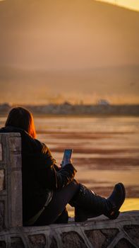 A woman sitting on a wall uses her smartphone at sunset by Beyşehir Lake, Türkiye.