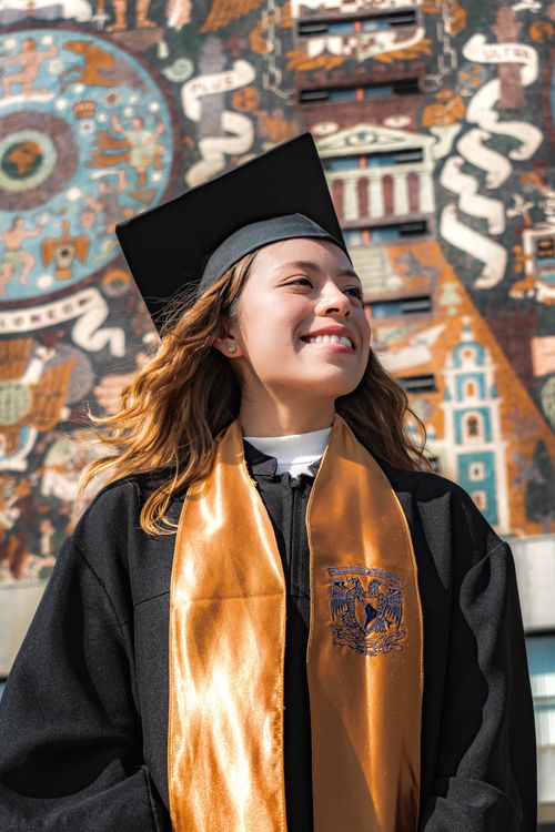 Free A proud young woman in a graduation gown smiles against a vibrant mural backdrop. Stock Photo