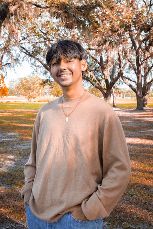Young Man in a Brown Sweater and Jeans Standing in a Park and Smiling 