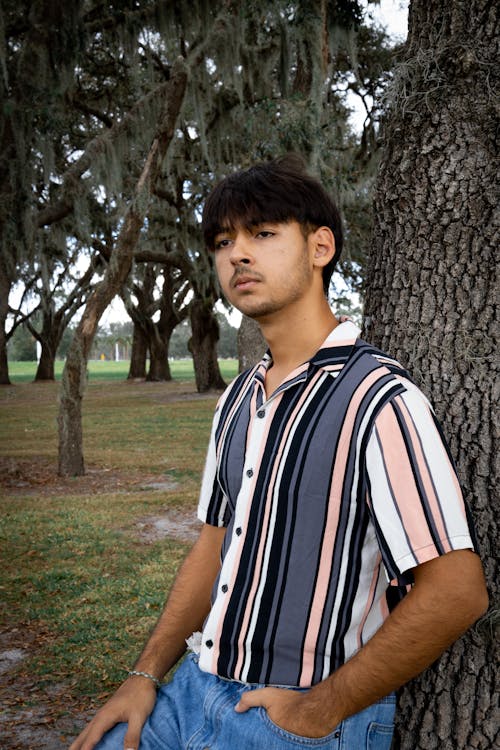 Young Man in a Striped Shirt and Jeans Standing in a Park 