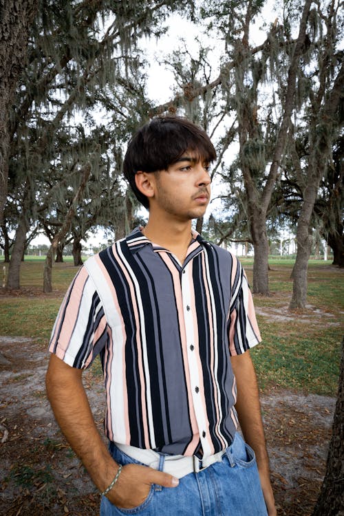 Young Man in a Striped Shirt and Jeans Standing in a Park 