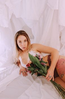 Elegant woman with long hair posing on white fabric, holding a bouquet of flowers.