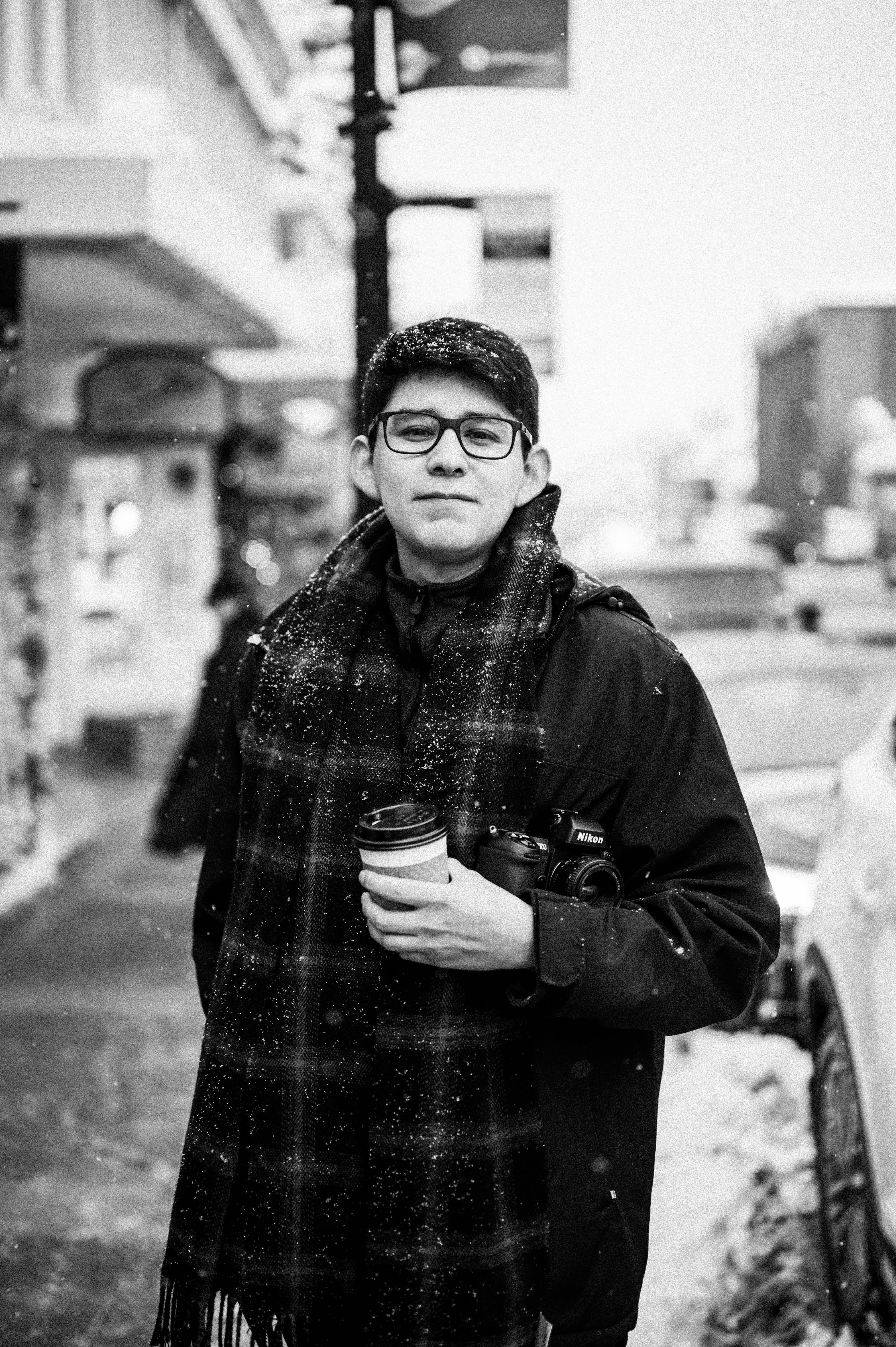 A man in winter clothing holding a coffee and camera on a snowy street.