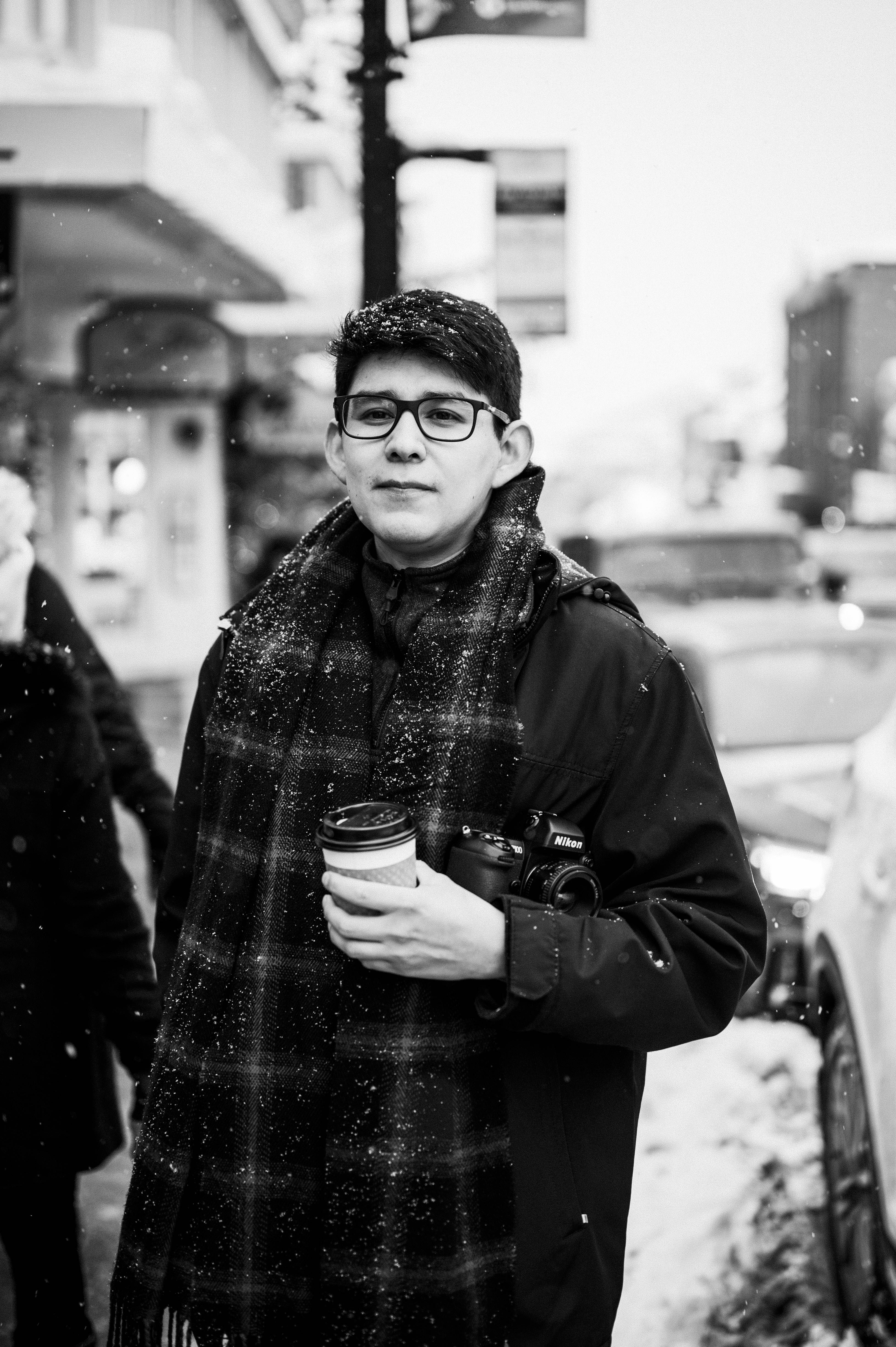 Black and white portrait of a photographer with coffee on a snowy urban street.