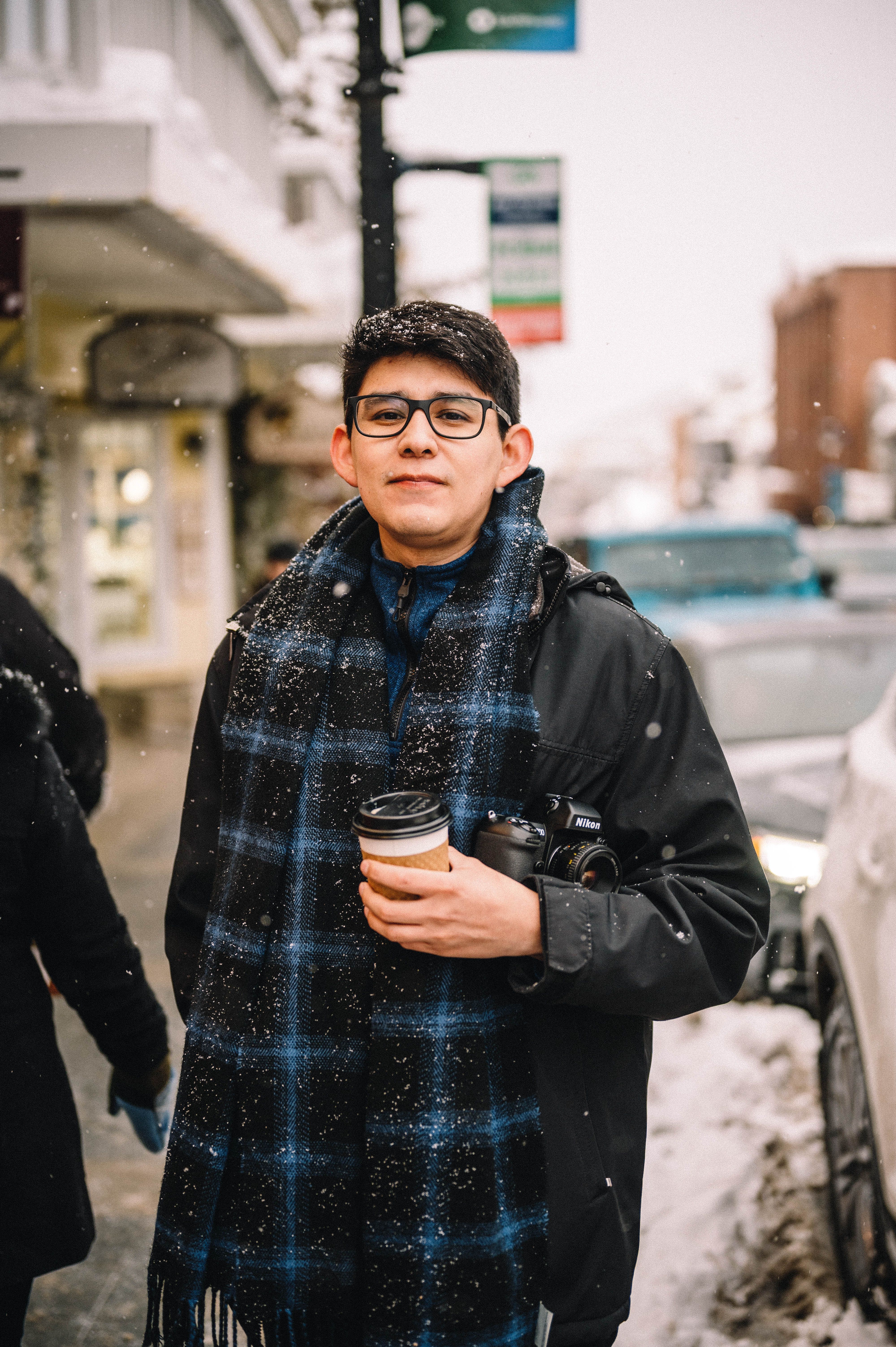 Man holding coffee and camera in a snowy urban setting, showcasing winter fashion and lifestyle.