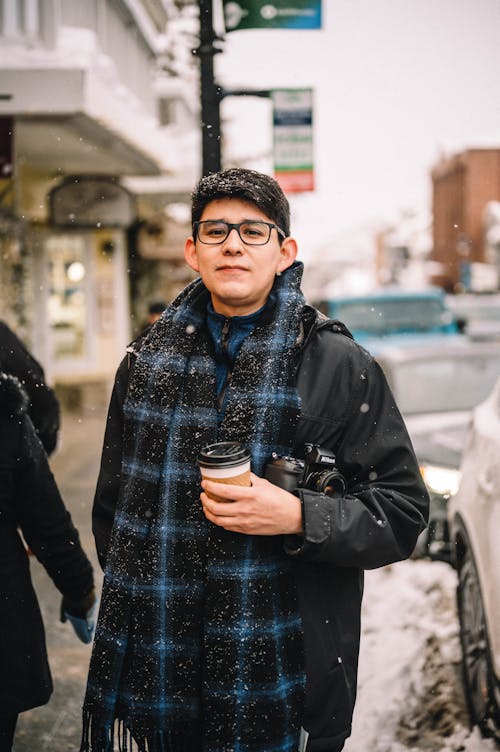 Passerby with a Cup of Coffee and a Camera on the Sidewalk Amidst the Falling Snow
