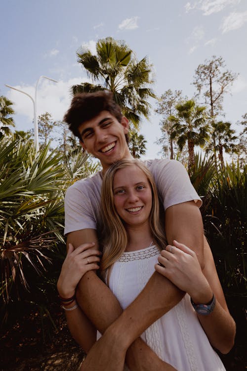 Free Smiling Young Man Standing Behind and Embracing a Woman in White Dress Stock Photo