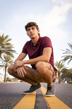Young man in casual attire squatting on a palm-lined street with a confident look.