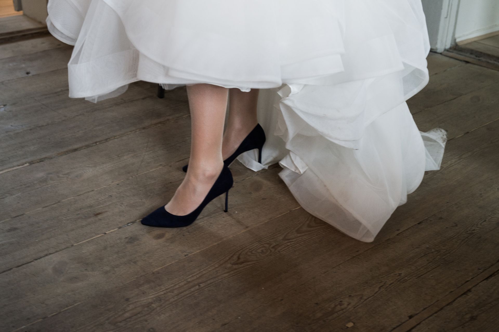 Close-up of a bride's feet in black heels under a white wedding dress on a wooden floor.