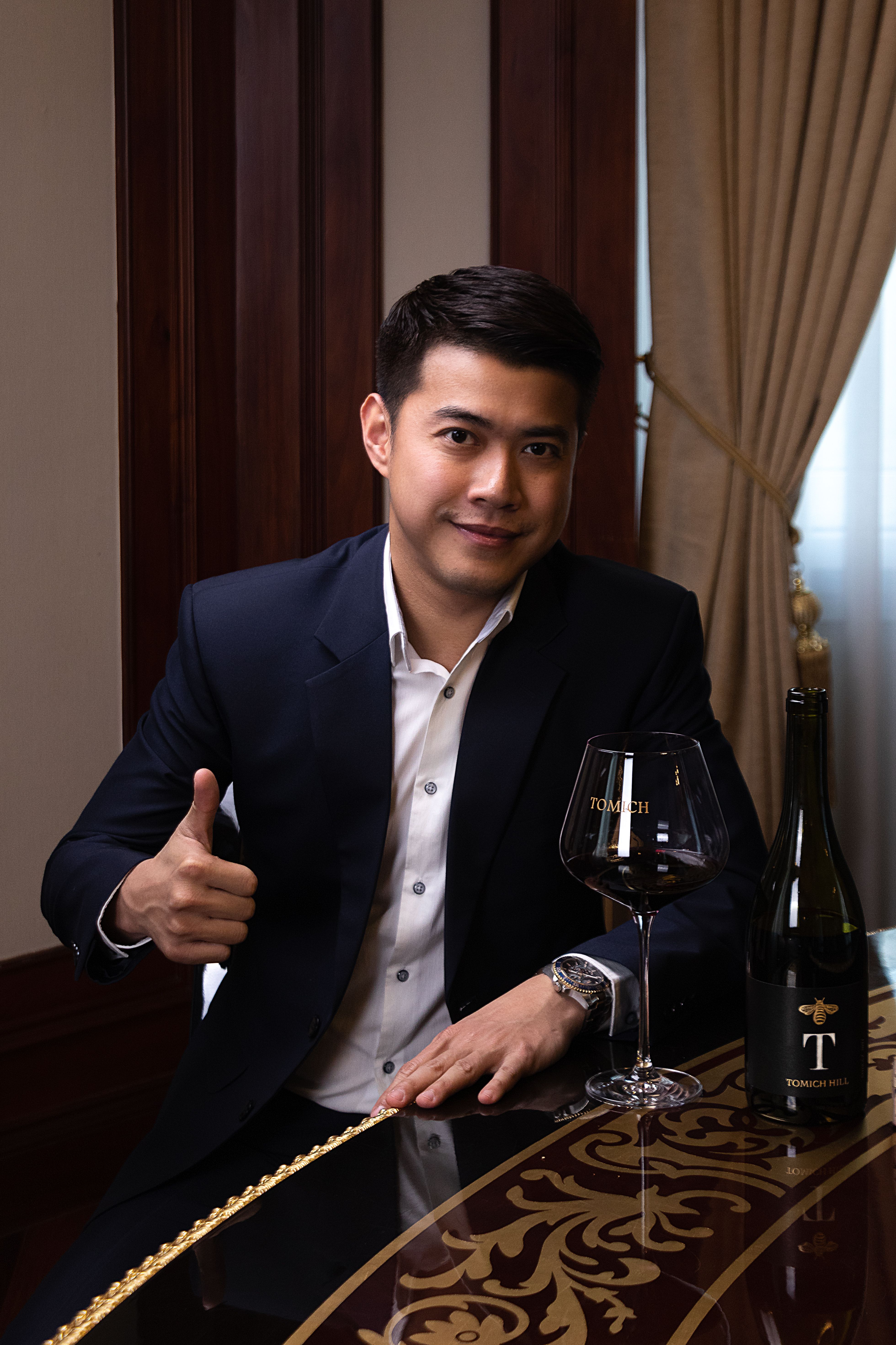 A confident man in a suit gives a thumbs up while enjoying a glass of wine indoors.
