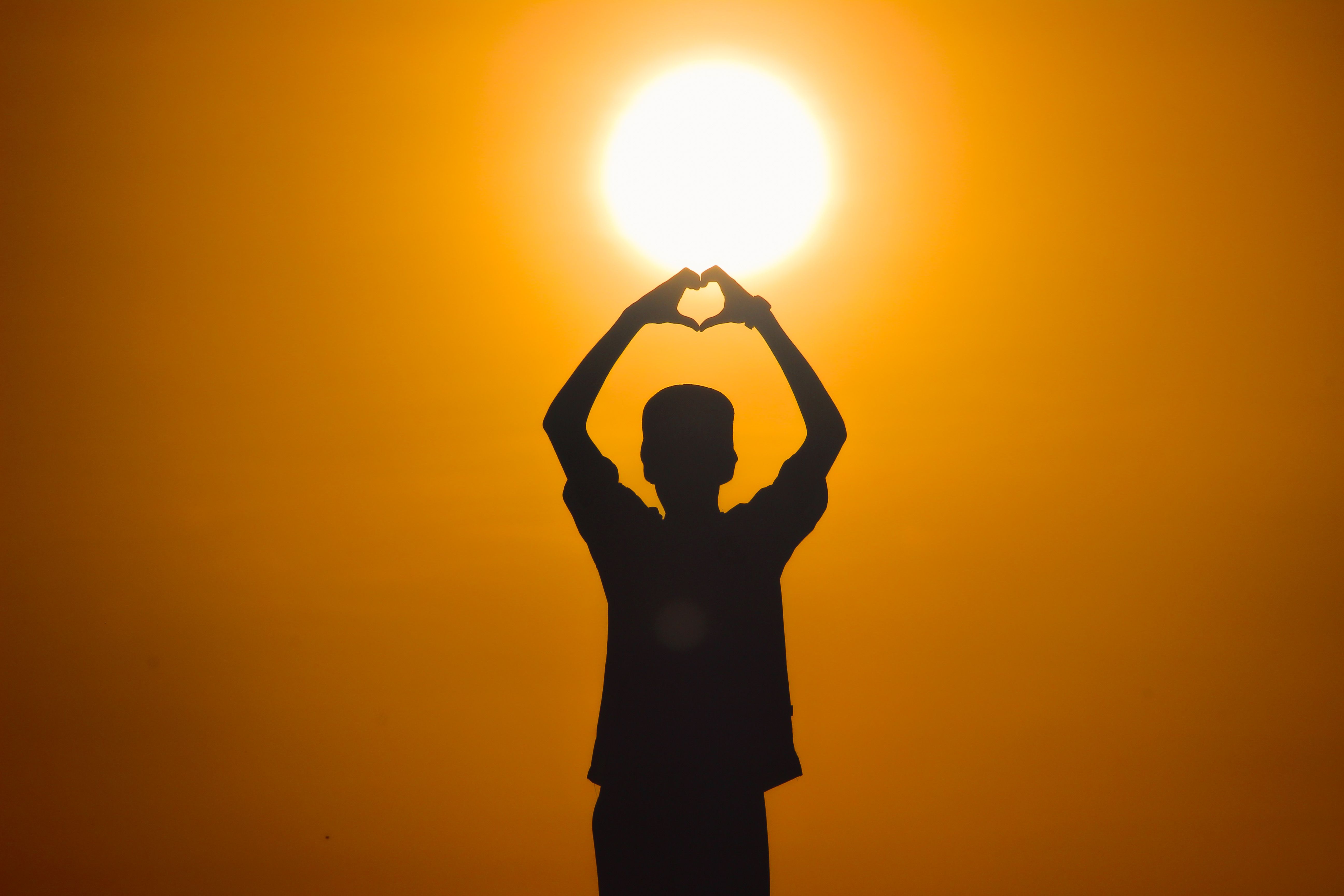 Silhouette of an individual forming a heart shape with hands against a bright sunset in Saudi Arabia.