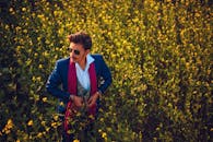 一位穿着优雅、身着蓝色西装、戴着太阳镜的男子，在白天于郁郁葱葱的花田中摆着姿势