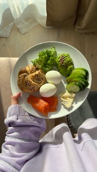 Plate of healthy breakfast including smoked salmon, eggs, and avocado held by a person indoors.