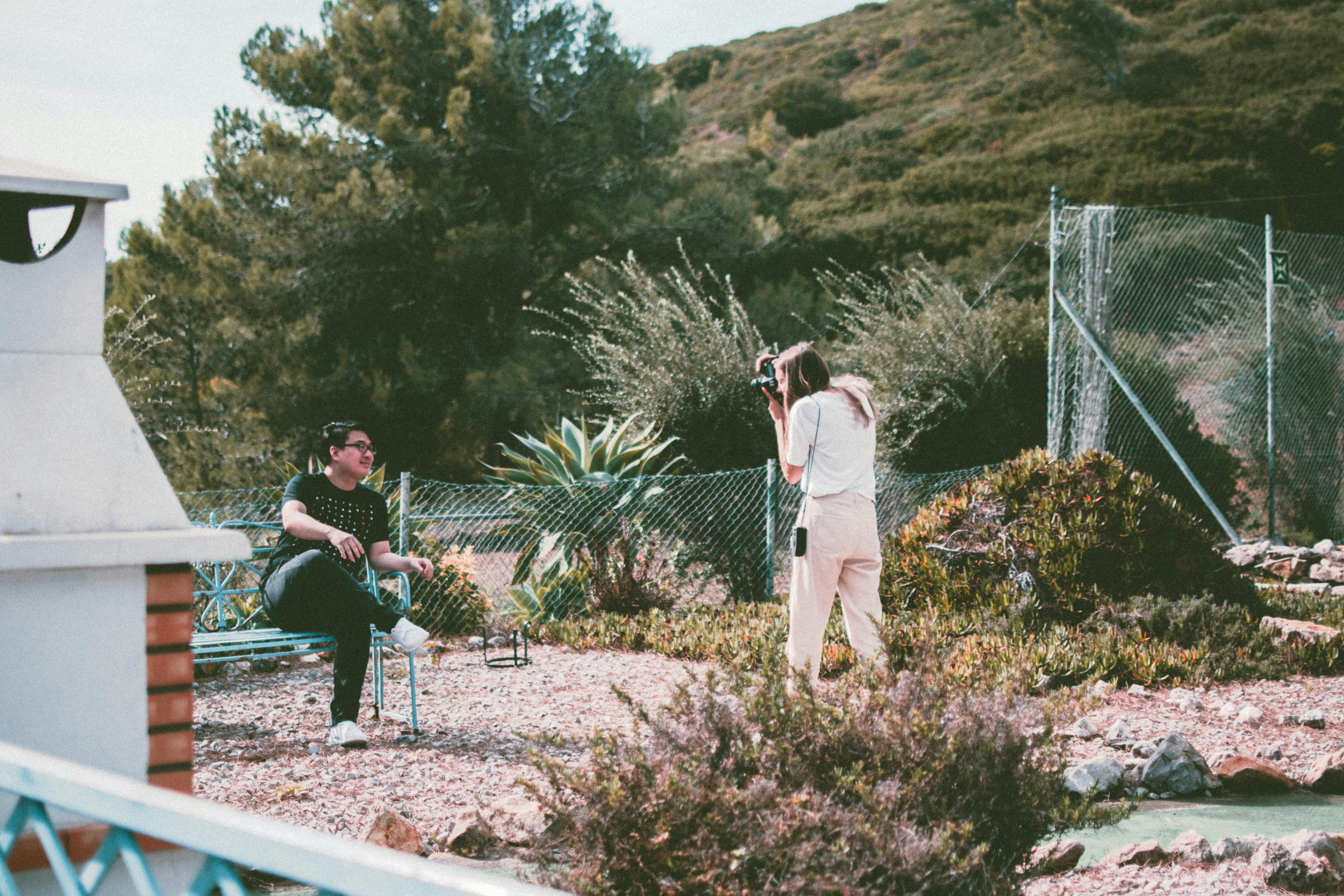 Man posing on a bench while a woman photographer takes pictures in a sunny garden.