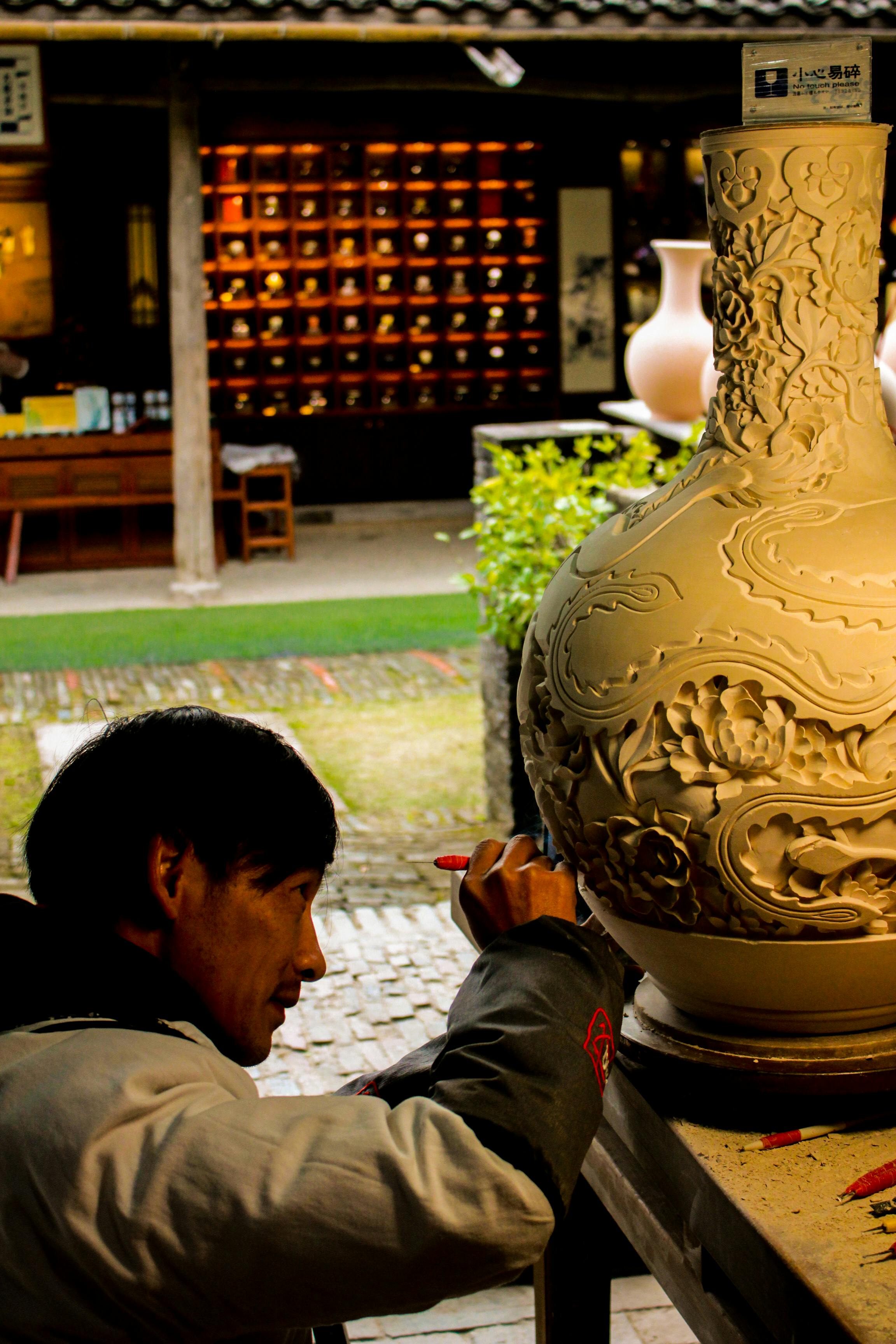 A skilled artisan intricately decorates a ceramic vase outdoors, showcasing craftsmanship.