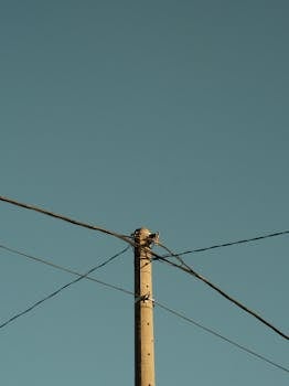 A minimalist vertical shot of an electrical pole with wires against a clear blue sky.