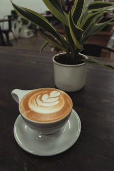 Close-up of a cappuccino with latte art beside a potted plant on a wooden table.