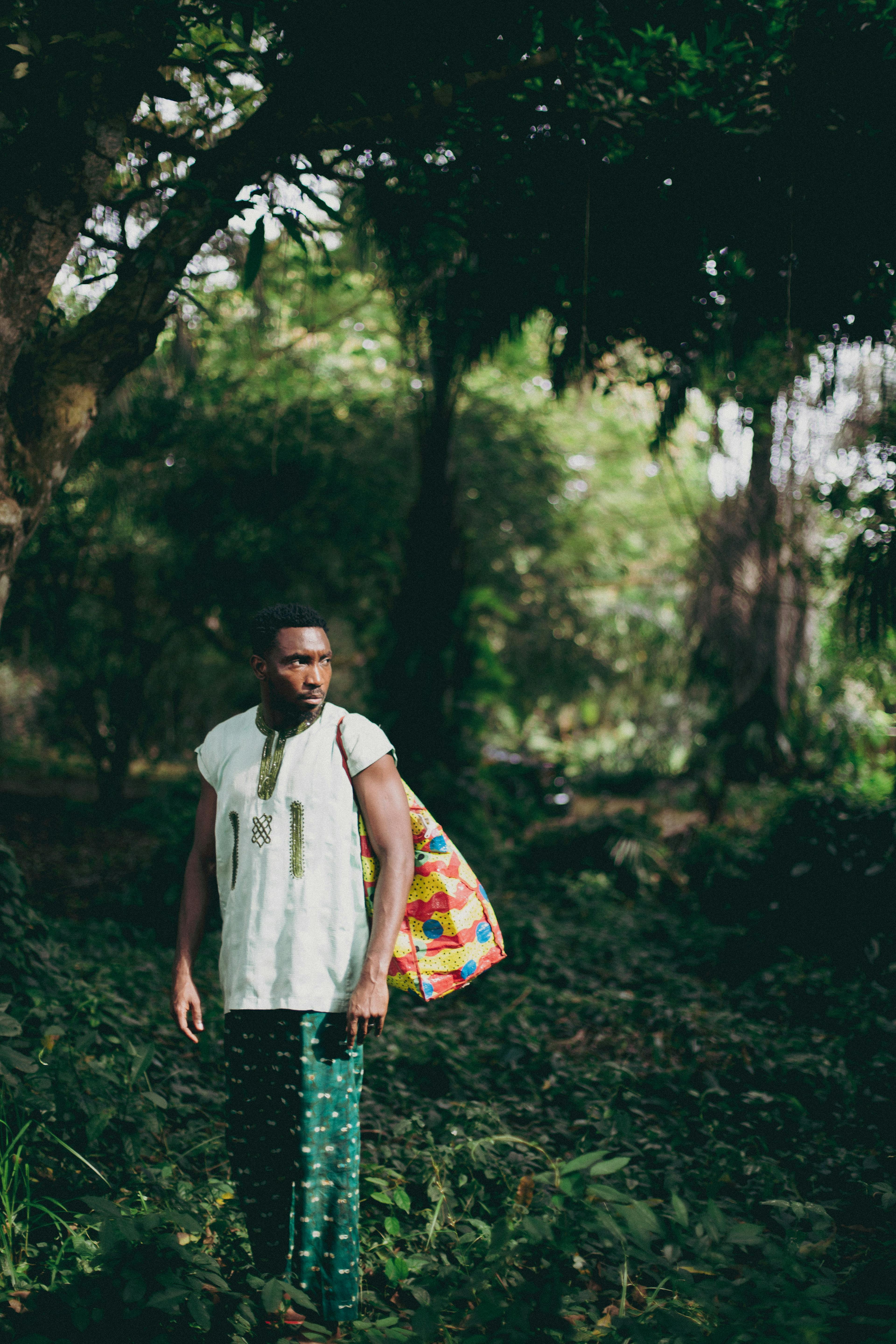 Portrait of a man in Lagos forest wearing traditional attire with a colorful bag, surrounded by lush greenery.