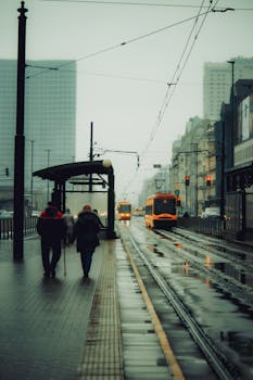 Two people walk along a rainy tram station, trams in the distance, creating a moody cityscape.