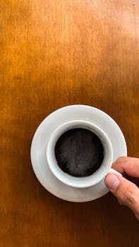 A hand holding a cup of black coffee on a wooden table, viewed from above.
