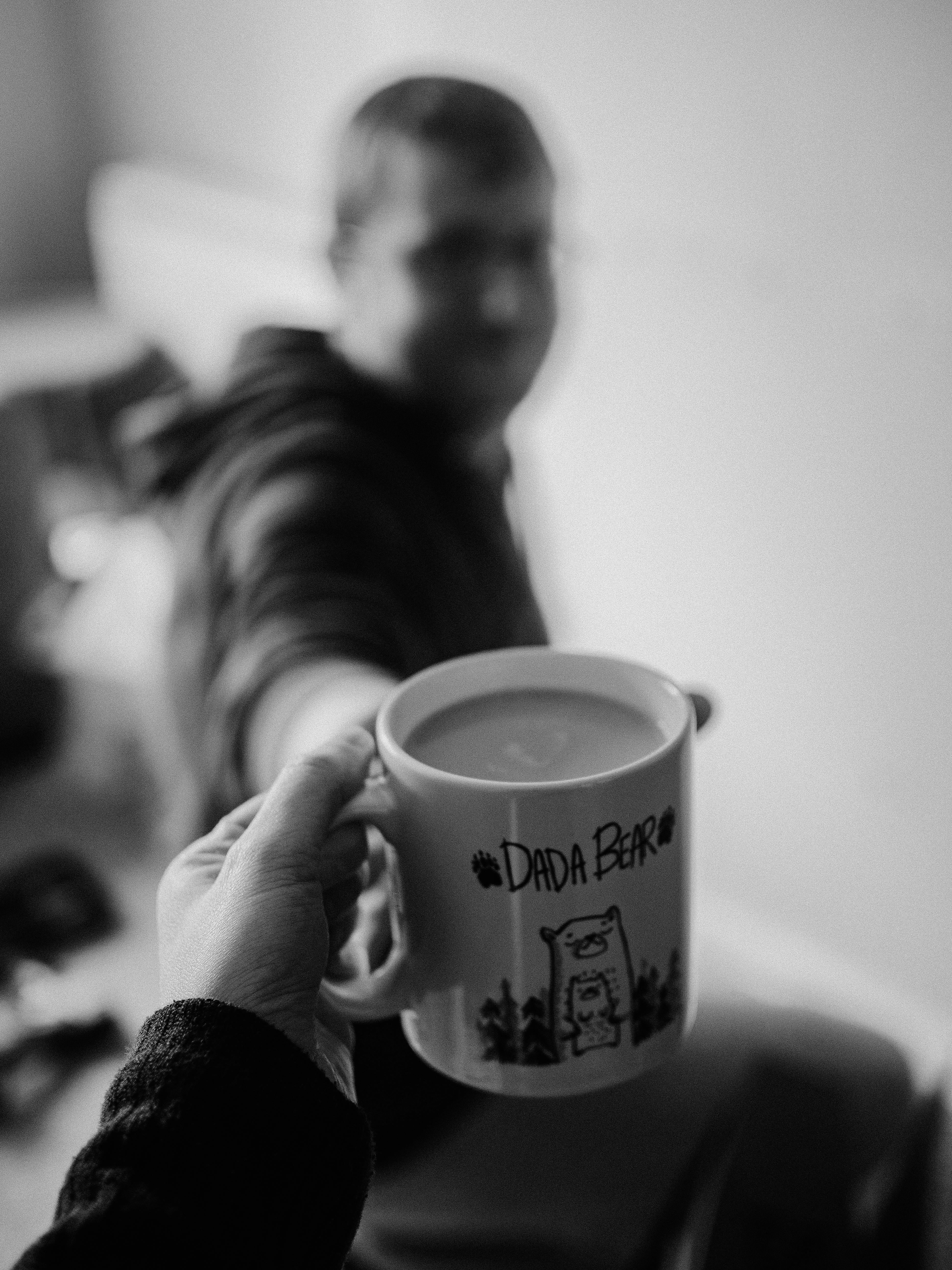 A person hands a 'Dada Bear' mug with a hot drink in a black and white setting.