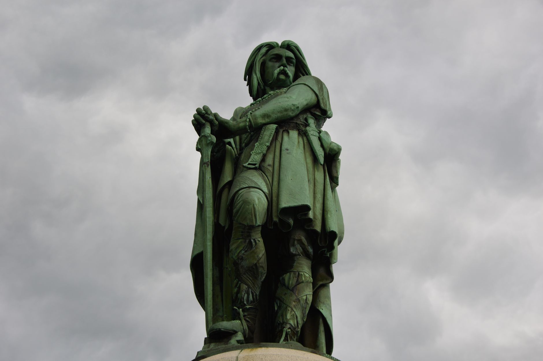 A powerful low-angle photograph of the Vercingetorix statue under a dramatic cloudy sky.